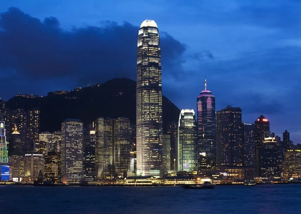 Horizonte de arranha-céus iluminados de Hong Kong ao anoitecer, visto do outro lado da baía, com a silhueta escura do morro atrás dos prédios