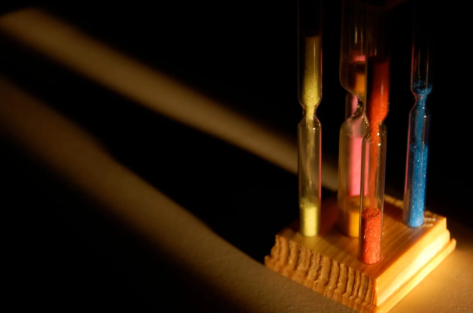 Four sand timers side by side in a wooden stand, each with a different amount of yellow, orange, red and blue sand left, lit from the side against a dark background