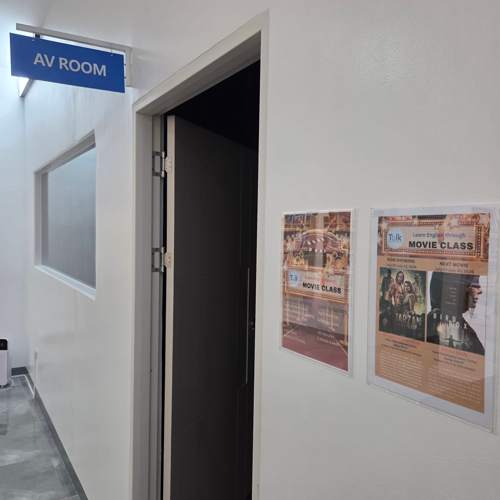 열린 문 위에 파란 AV ROOM 표지판이 걸린 어학원 복도, 벽에는 무비 클래스 포스터 2장