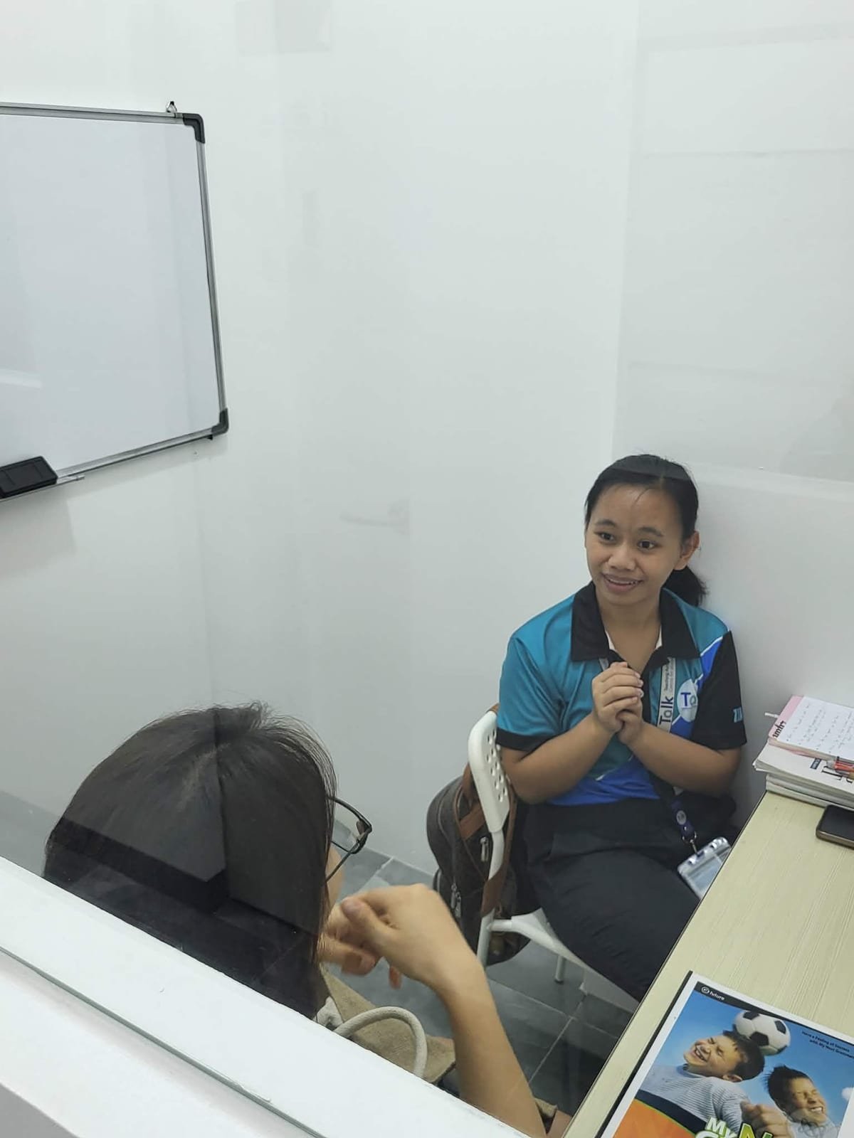 A one-on-one lesson in a booth-style classroom, teacher and student facing each other across a desk